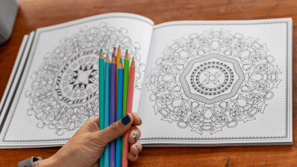 Pessoa segurando lápis de cores, com livro para colorir ao fundo.